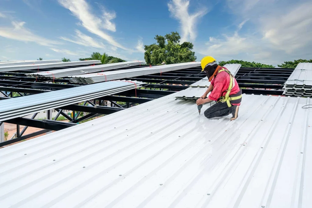 A worker from Precision Construction Systems kneels in a safety harness on a metal roof, meticulously installing white corrugated panels against a backdrop of blue sky and scattered clouds.